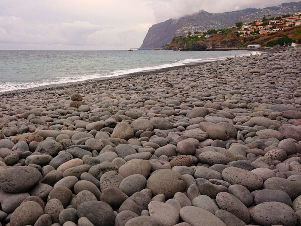 Ein Strand mit grossen Kieselsteinen bei bedecktem Himmel. Im Hintergrund ist eine Klippe zu sehen.